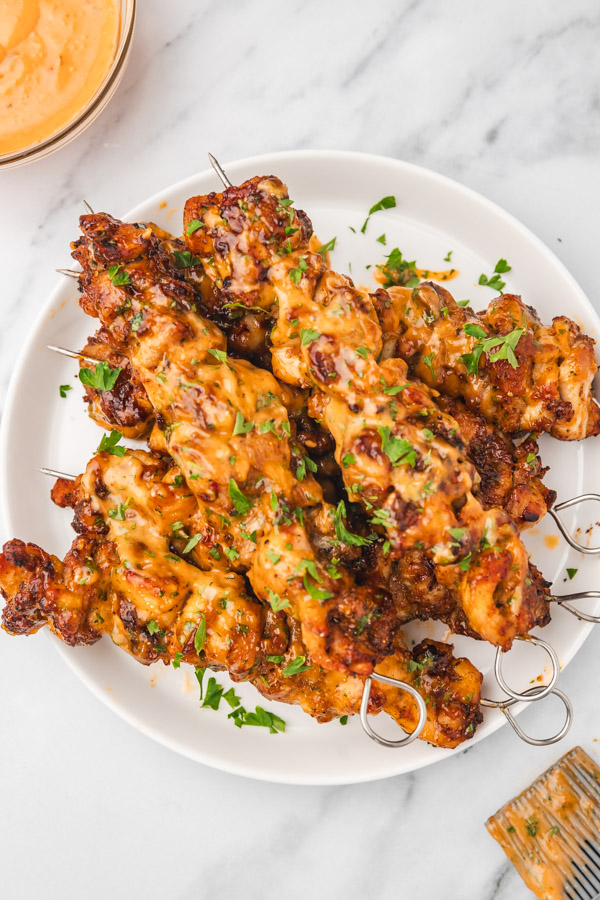 chicken skewers stacked on a white plate placed beside a small pot of sippings sauce.